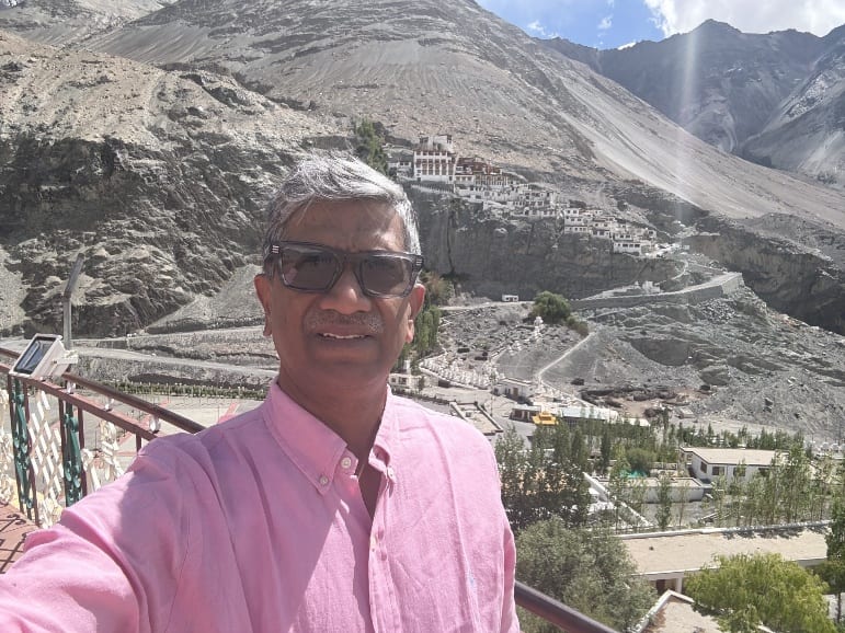 George Kovoor in a pink shirt on a terrace in Nubra Valley, Ladakh, with a hillside monastery and bare mountains behind him.