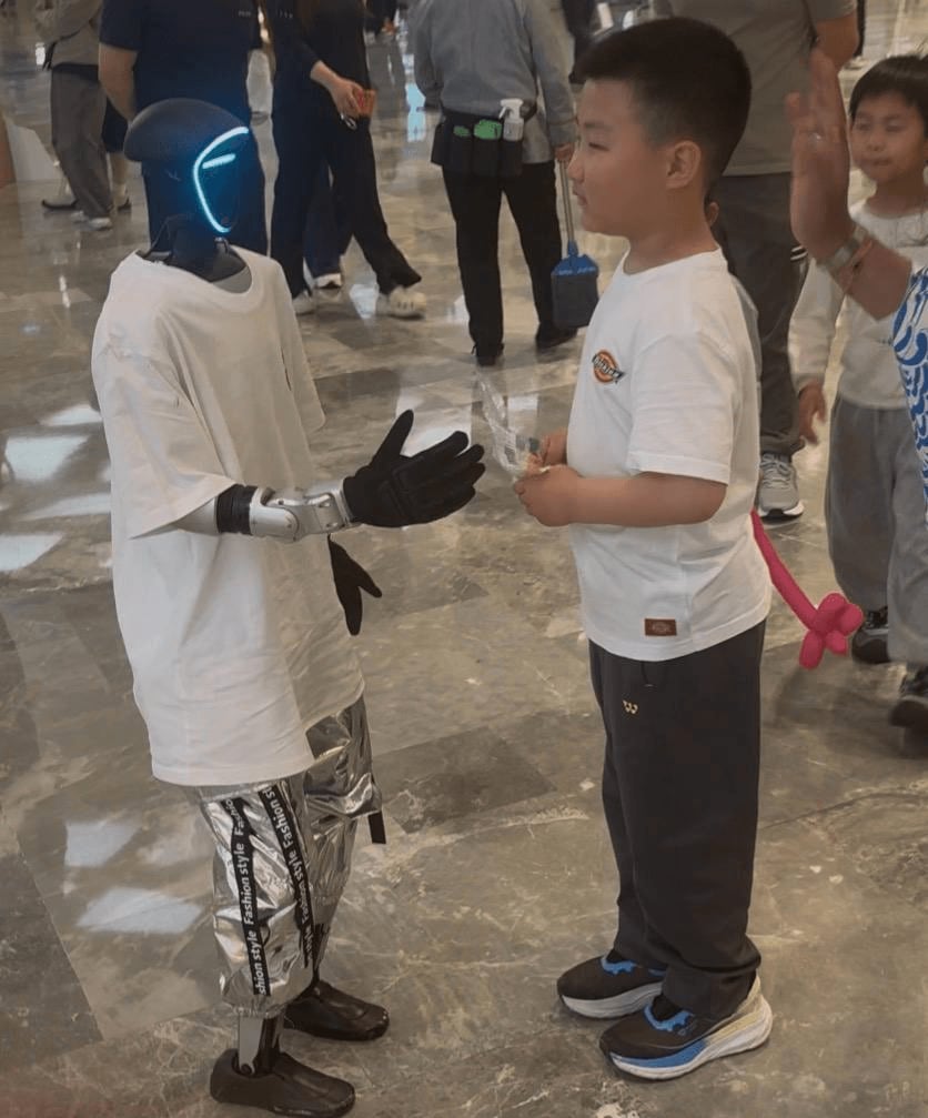 A humanoid robot in a promo shirt shaking hands with a young boy in a mall.